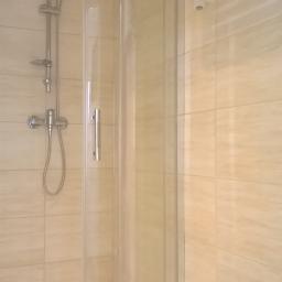 Norbert - Narożna kabina prysznicowa z transparentnymi drzwiami, chromowana bateria i deszczownica na tle beżowych, kwadratowych płytek oraz biały grzejnik drabinkowy.