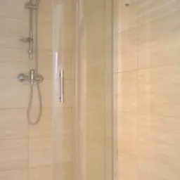 Narożna kabina prysznicowa z transparentnymi drzwiami, chromowana bateria i deszczownica na tle beżowych, kwadratowych płytek oraz biały grzejnik drabinkowy.