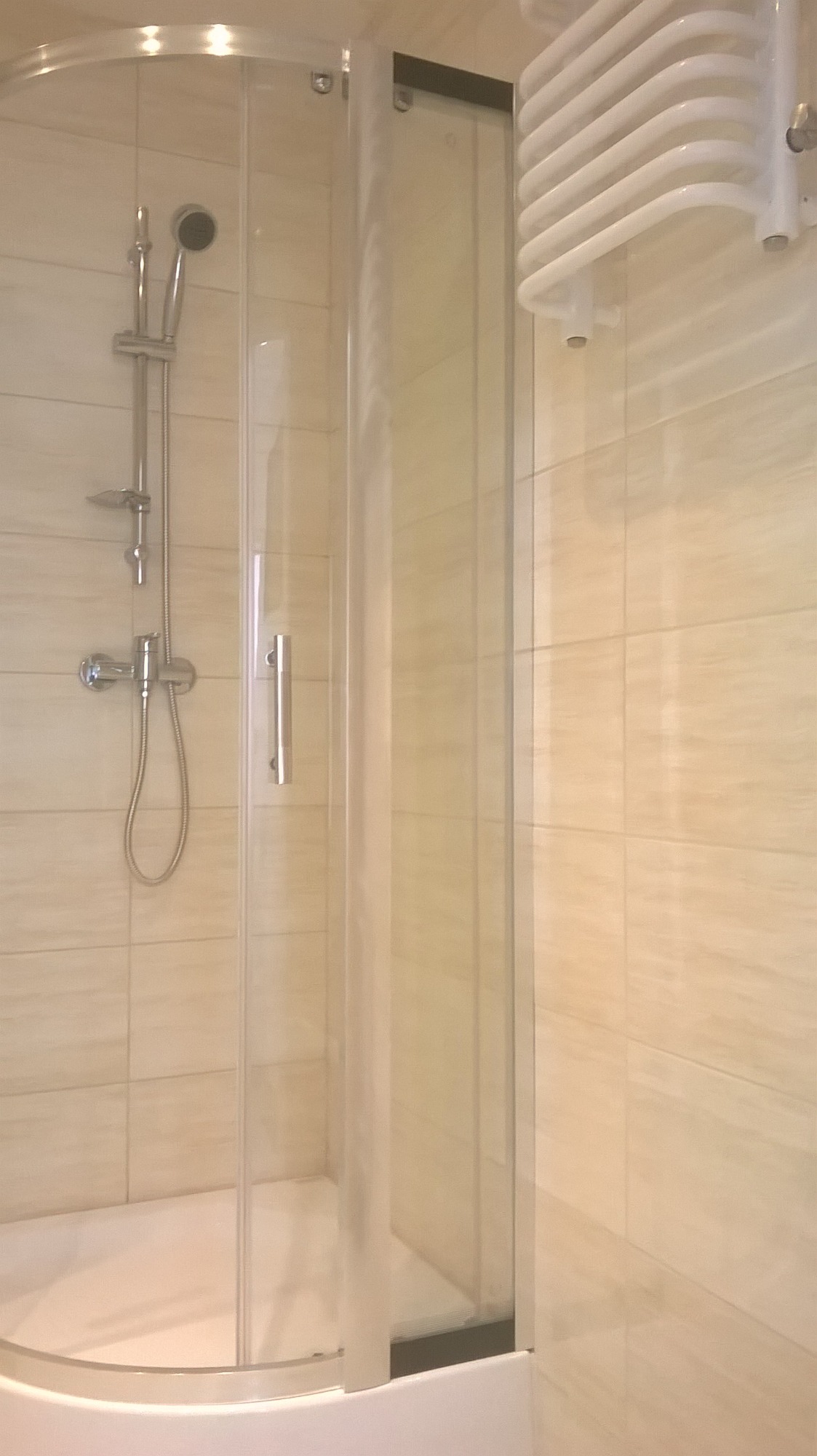 Narożna kabina prysznicowa z transparentnymi drzwiami, chromowana bateria i deszczownica na tle beżowych, kwadratowych płytek oraz biały grzejnik drabinkowy.