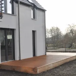 Nowoczesny, brązowy taras przylegający do szarego domu z czarną stolarką okienną, widoczny klimatyzator na ścianie, tło stanowią drzewa bez liści i ogrodzenie.