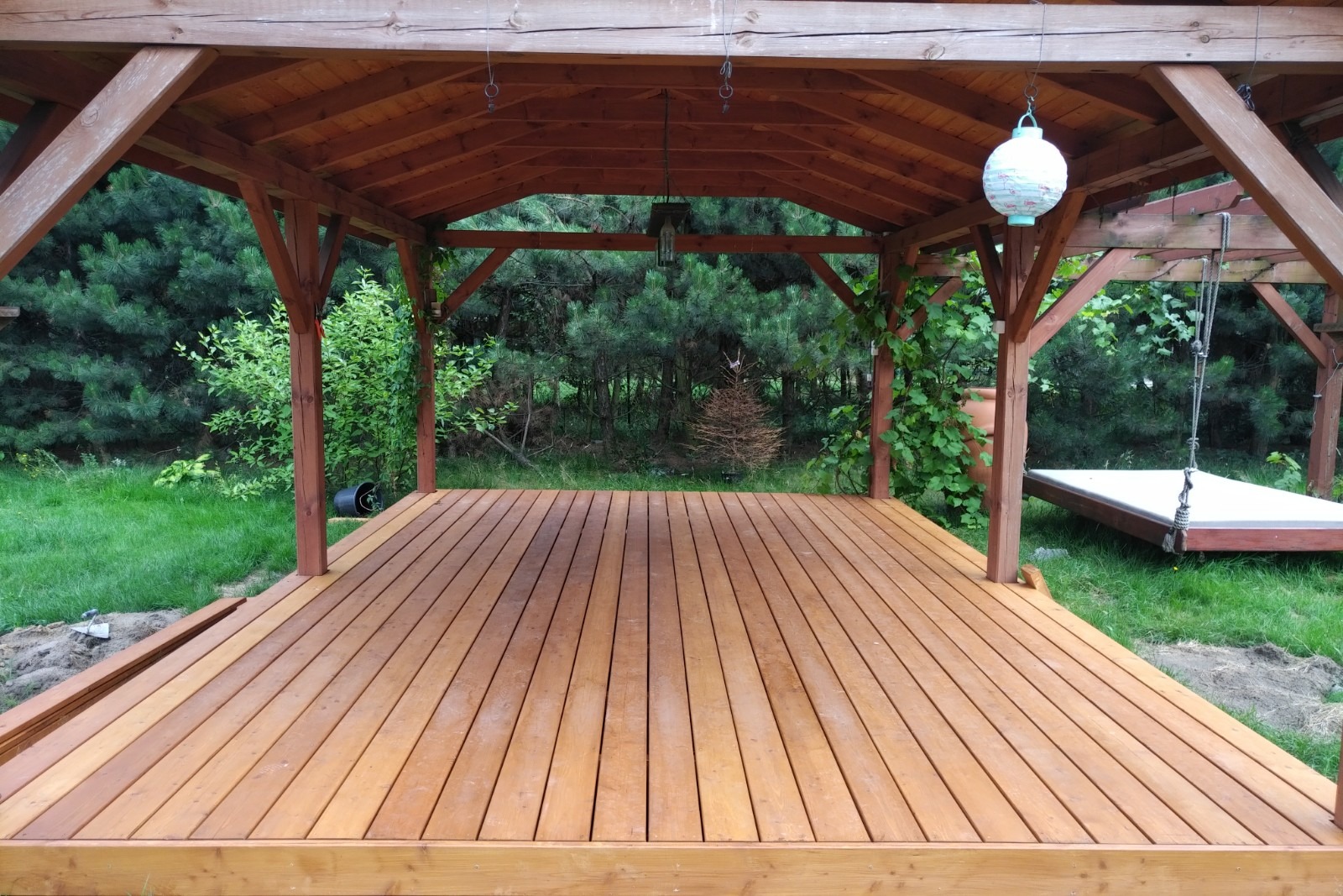Zadaszony taras z drewna z widokiem na zieleń, lampa papierowa zwisająca z sufitu, huśtawka na drugim planie.