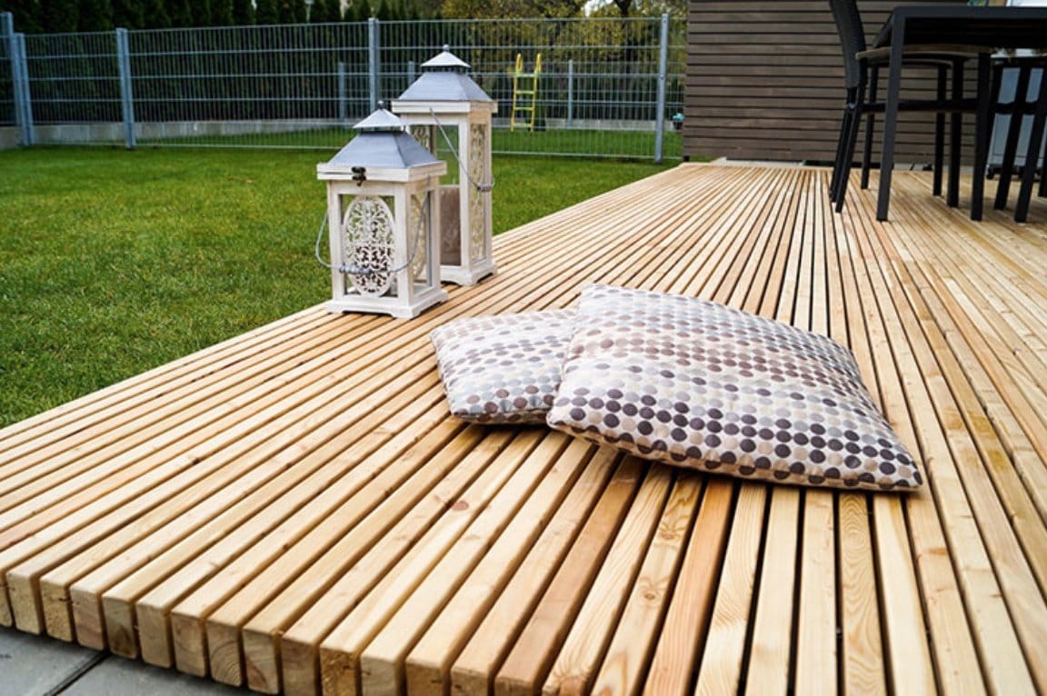Nowoczesny taras z desek ułożonych równolegle, z dwoma dekoracyjnymi lampionami i poduszkami w kropki, na tle zadbanego trawnika i metalowego ogrodzenia.