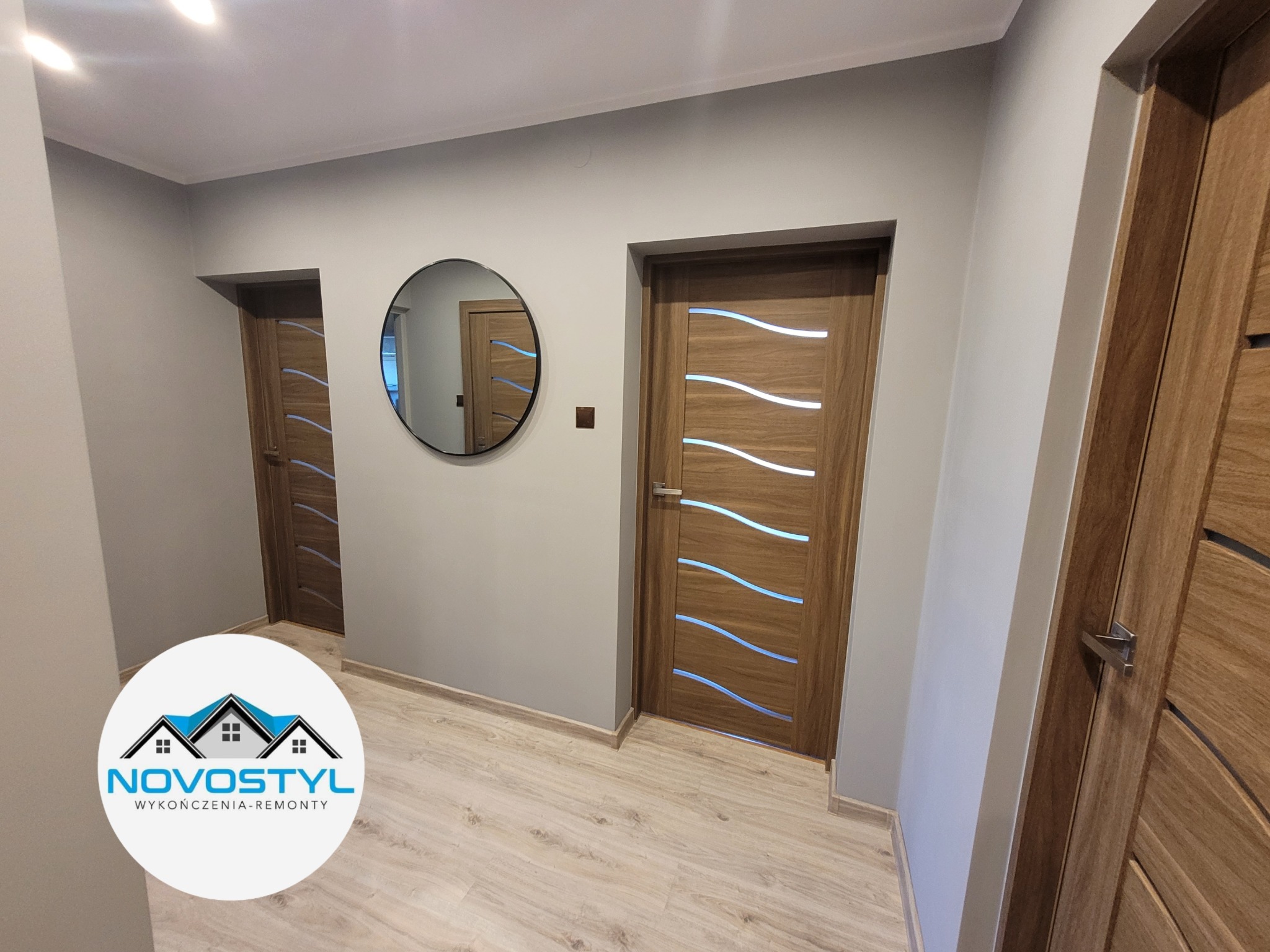 Korytarz po remoncie z dwiema drewnianymi drzwiami z poziomymi, falistymi przeszkleniami, okrągłe lustro na ścianie, podłoga wyłożona panelami, logo firmy na pierwszym planie.