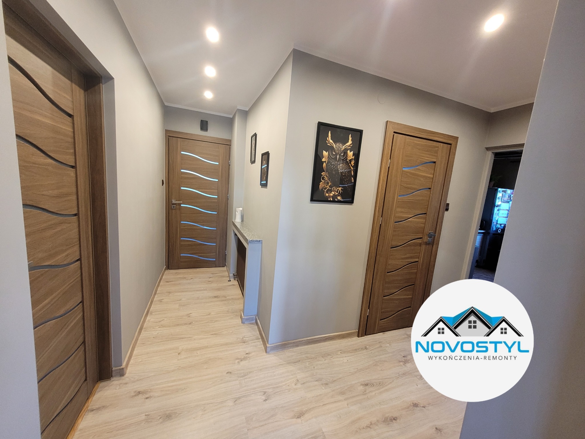 Korytarz po remoncie z trzema drewnianymi drzwiami z dekoracyjnymi wstawkami, panelami podłogowymi, szarą ścianą z obrazami i punktowym oświetleniem sufitowym.