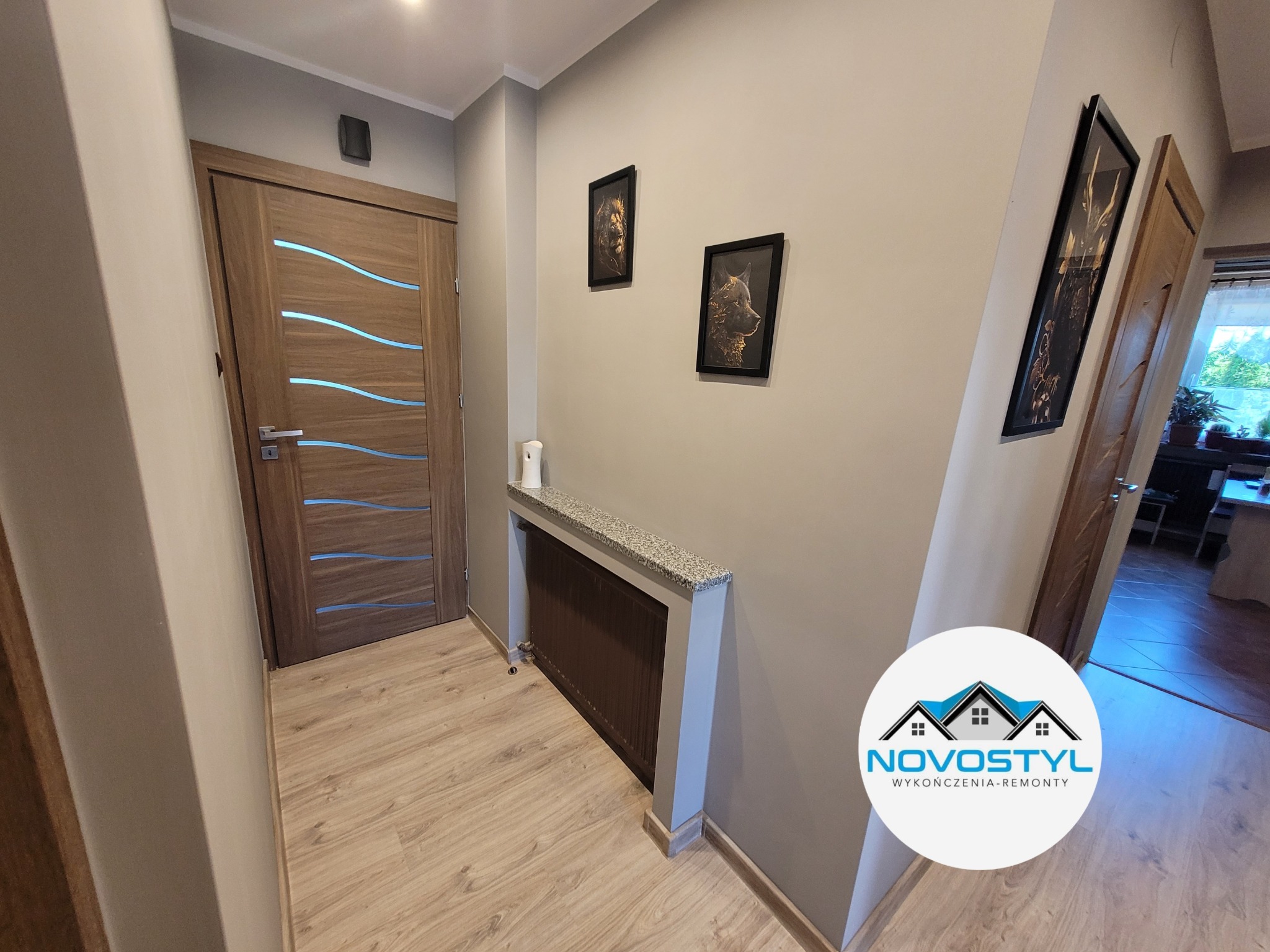Korytarz po remoncie z drewnianymi drzwiami z poziomymi przeszkleniami, panelami podłogowymi, zabudowanym grzejnikiem z blatem z lastryko i obrazami na ścianie.