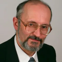 Portret mężczyzny w okularach i garniturze, z brodą i wąsami, na jasnym tle.