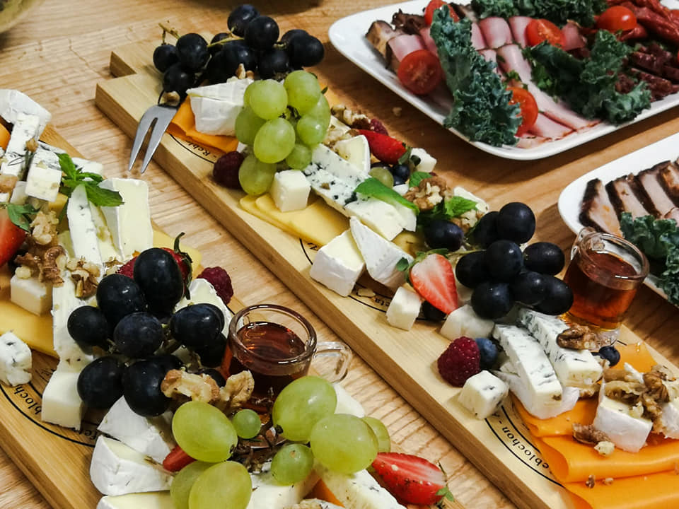 Dwie drewniane deski z serami brie, pleśniowymi, żółtymi, winogronami, truskawkami, malinami, orzechami włoskimi i miodem w małych szklankach, na tle talerzy z wędlinami i pomidorami.