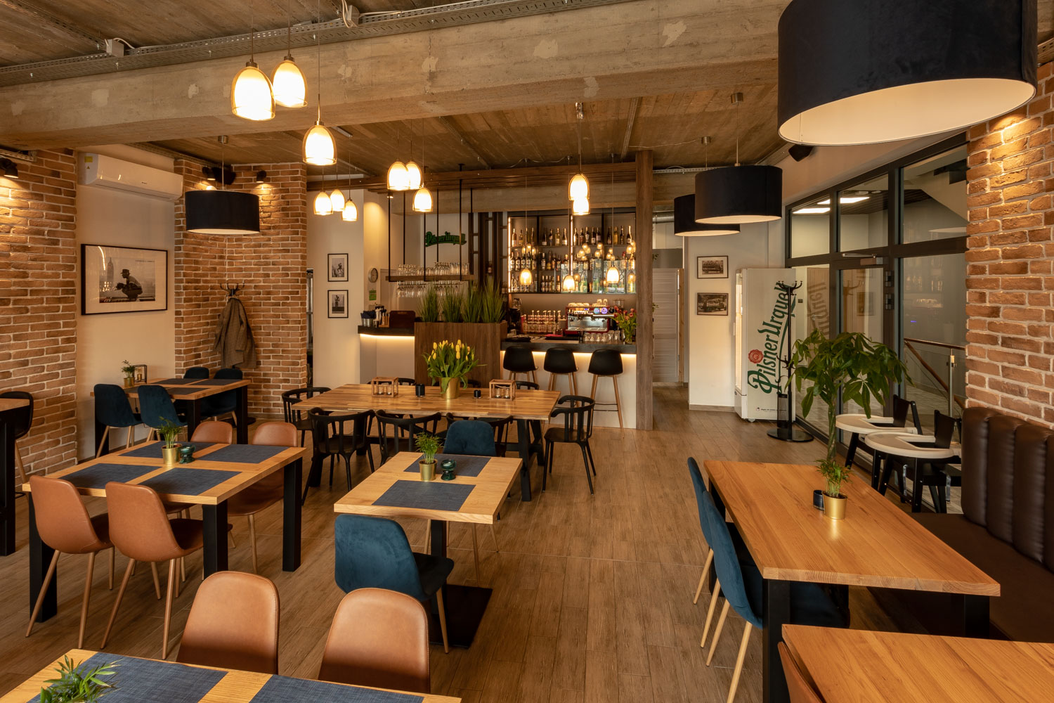 Wnętrze restauracji z drewnianymi stołami, krzesłami w odcieniach brązu i granatu, bar z oświetleniem punktowym i lodówka Pilsner Urquell w tle.