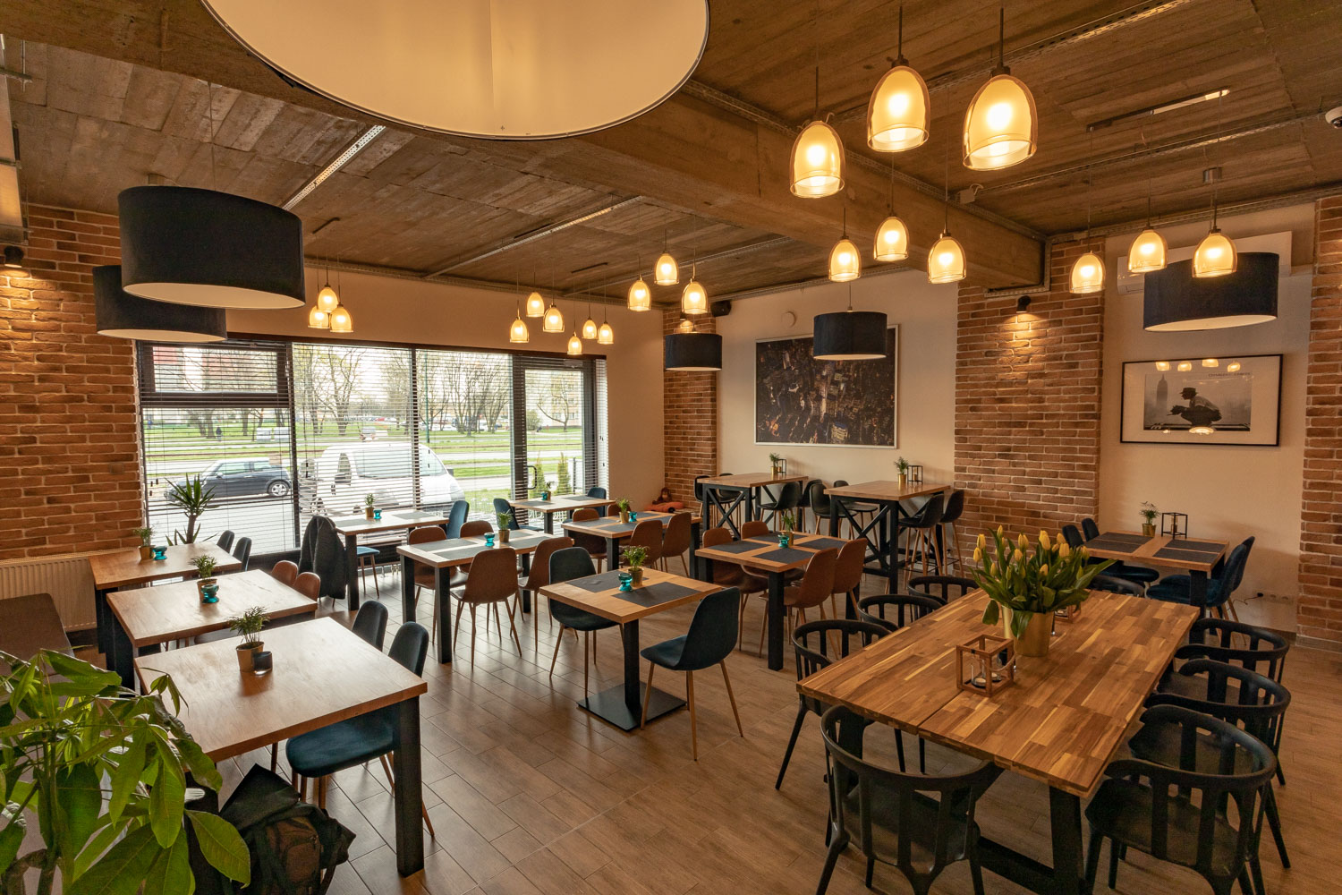 Jasne wnętrze restauracji z drewnianymi stołami, różnymi krzesłami, ceglanymi ścianami, licznymi lampami wiszącymi i widokiem na ulicę przez duże okno.