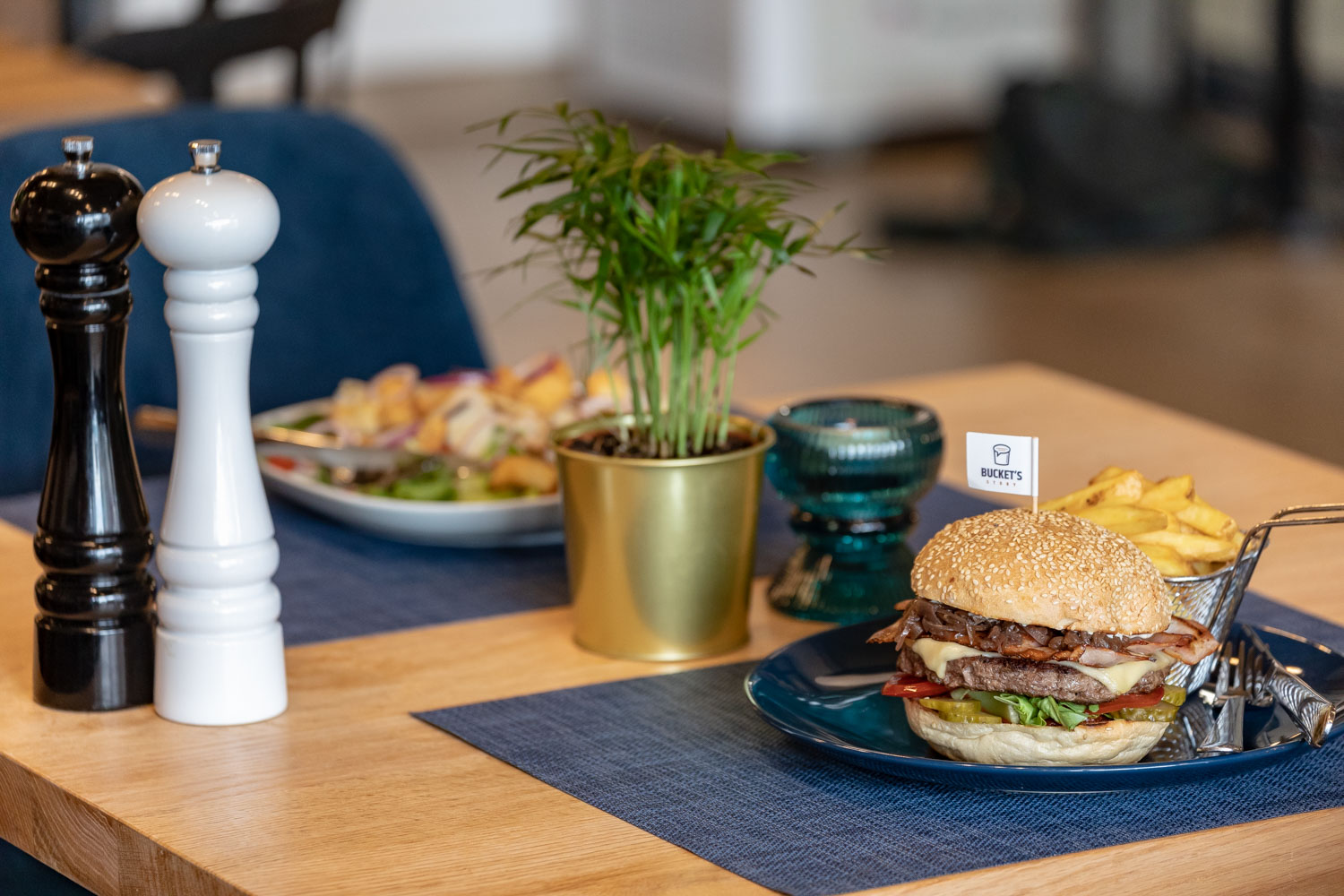 Burger z bekonem, serem i warzywami na niebieskim talerzu, frytki w metalowym koszyku, sałatka, przyprawniki i roślina w złotym garnku na drewnianym stole z niebieską podkładką.