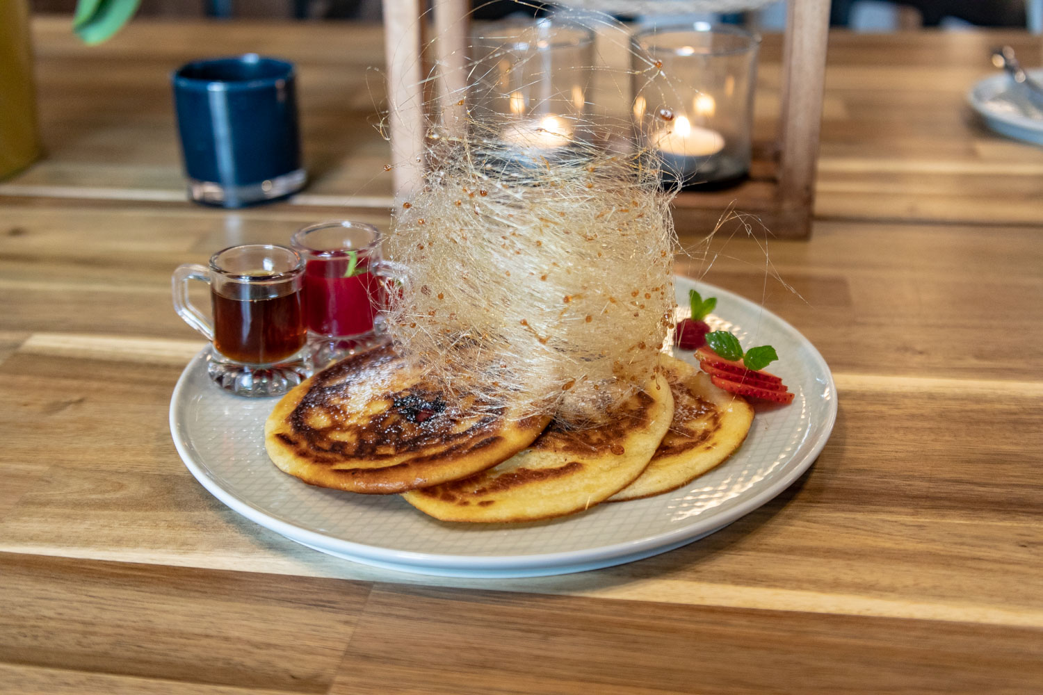 Trzy puszyste naleśniki posypane cukrem pudrem, udekorowane karmelową pajęczyną, malinami, plasterkami truskawek i listkami mięty, podane z dwoma małymi dzbanuszkami z syropem klonowym i sosem...