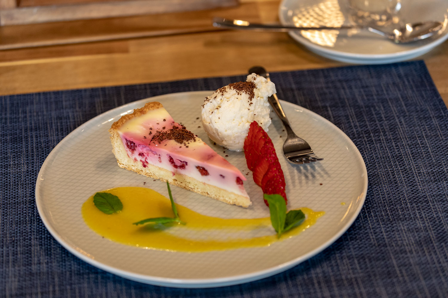 Porcja tarty z owocami i sosem mango, podana z gałką lodów i świeżą truskawką, udekorowana listkami mięty i czekoladą wiórkową na talerzu z delikatnym wzorem.