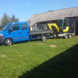 Niebieski samochód dostawczy z platformą przewozi na przyczepie żółto-zieloną mini koparkę marki Yanmar, zaparkowany na trawniku przed szarym, drewnianym budynkiem gospodarczym.