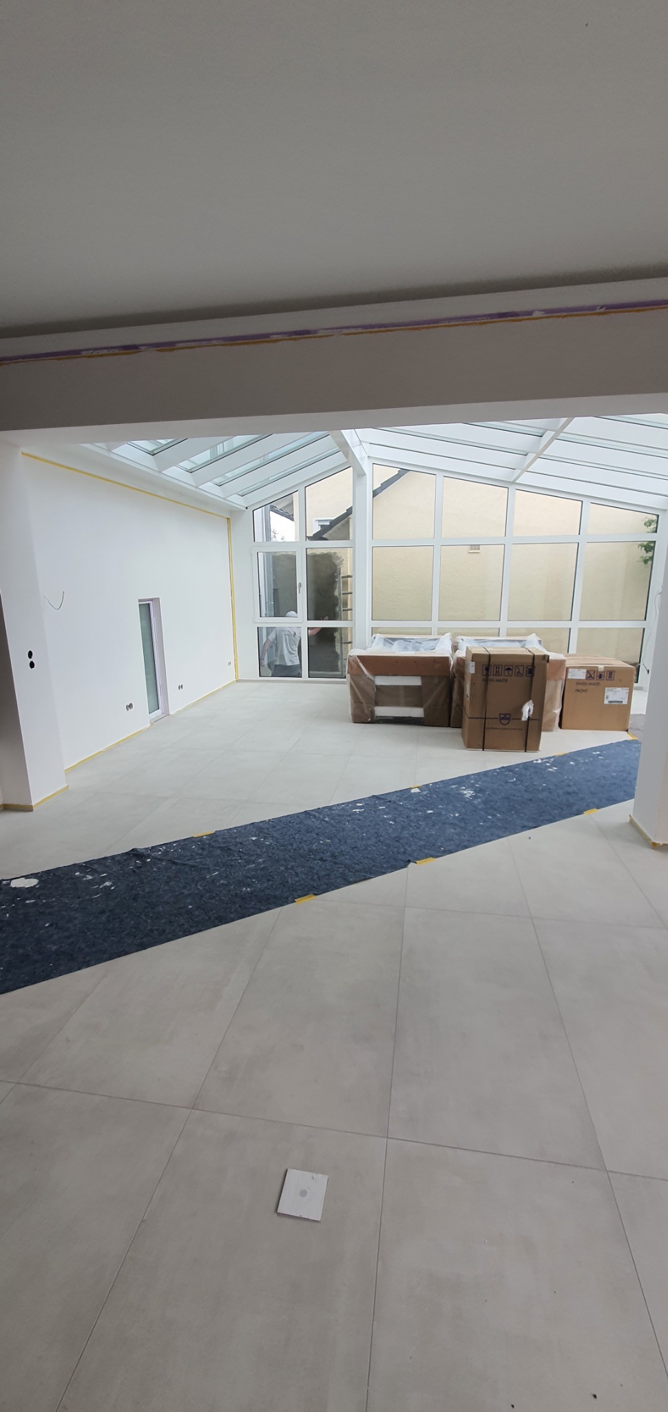 Jasne wnętrze w trakcie wykańczania: świeżo pomalowane ściany z taśmą malarską, zabezpieczona podłoga, przeszklona weranda z kartonami w tle.