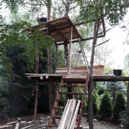 Konstrukcja drewnianego domku na drzewie w trakcie budowy, widoczne deski, drabina i osoba na górze, otoczenie leśne.