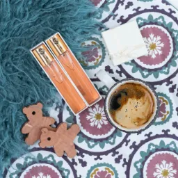 Dwa flakoniki perfum w pudełkach obok filiżanki kawy, prezentu i drewnianych zabawek na wzorzystej tkaninie i turkusowym futerku.