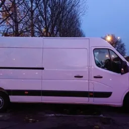 Biały samochód dostawczy Renault Master zaparkowany na poboczu w jesienny wieczór, widoczny budynek mieszkalny w tle.