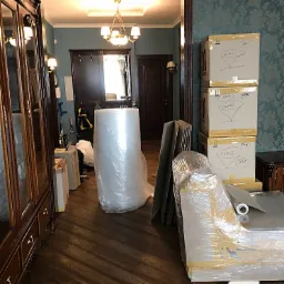 Wnętrze mieszkania z przygotowanymi do transportu przedmiotami: kartony z napisem 'GLASS', meble owinięte folią bąbelkową i stretch, panele zabezpieczone kartonem, widoczny parkiet i drzwi w tle.