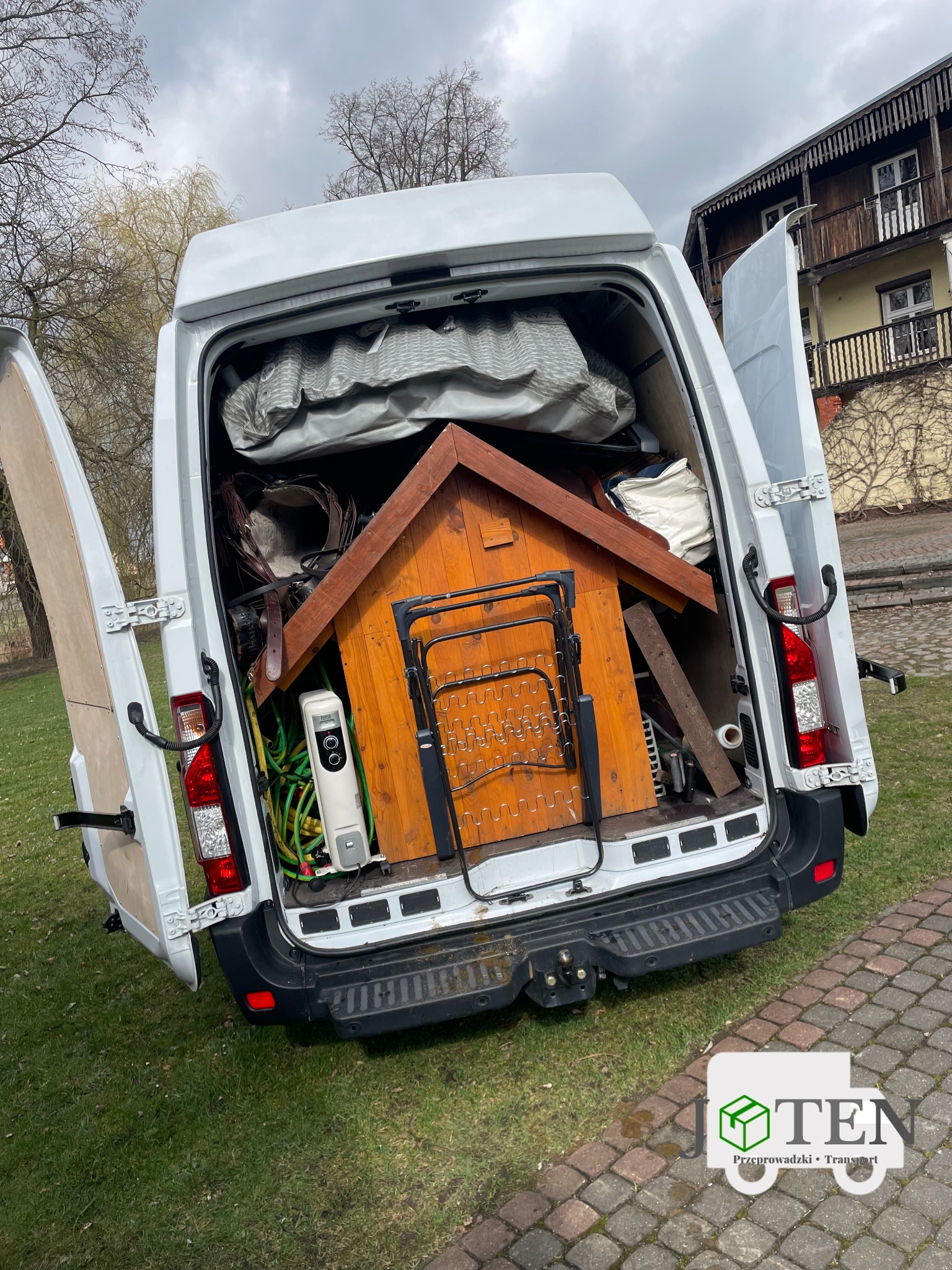 Otwarty bagażnik białego busa załadowany różnorodnymi przedmiotami, w tym budą dla psa, grzejnikiem elektrycznym, materacem i innymi pakunkami, na tle trawnika i budynku mieszkalnego.