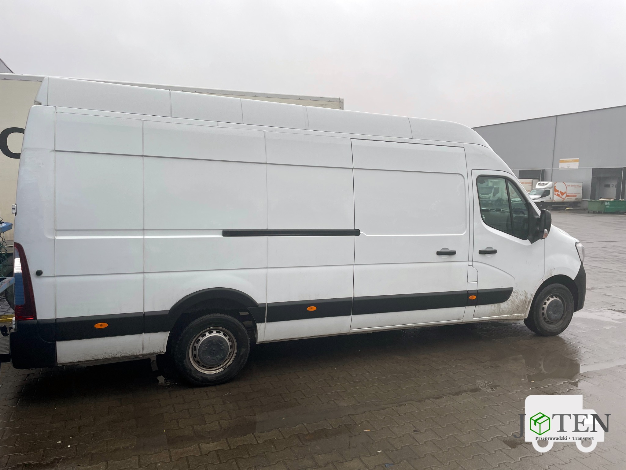 Biały samochód dostawczy Renault Master na mokrej kostce brukowej, widoczny z boku, z logo firmy transportowej w prawym dolnym rogu, na tle szarego budynku magazynowego i pochmurnego nieba.