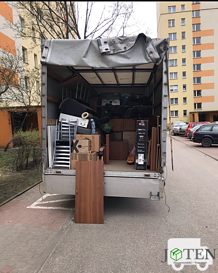 Otwarty tył ciężarówki załadowanej meblami i sprzętami podczas przeprowadzki, z widoczną częścią budynku mieszkalnego w tle, oraz brązowe drzwi opierające się o tył ciężarówki.