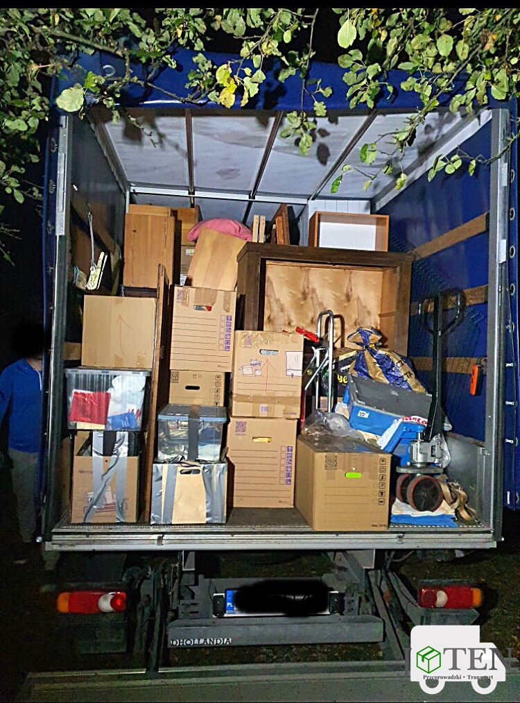 Wnętrze skrzyni ładunkowej wypełnione kartonami, meblami i sprzętem do transportu, widoczne gałęzie drzewa nad skrzynią, osoba w niebieskiej bluzie po lewej stronie.