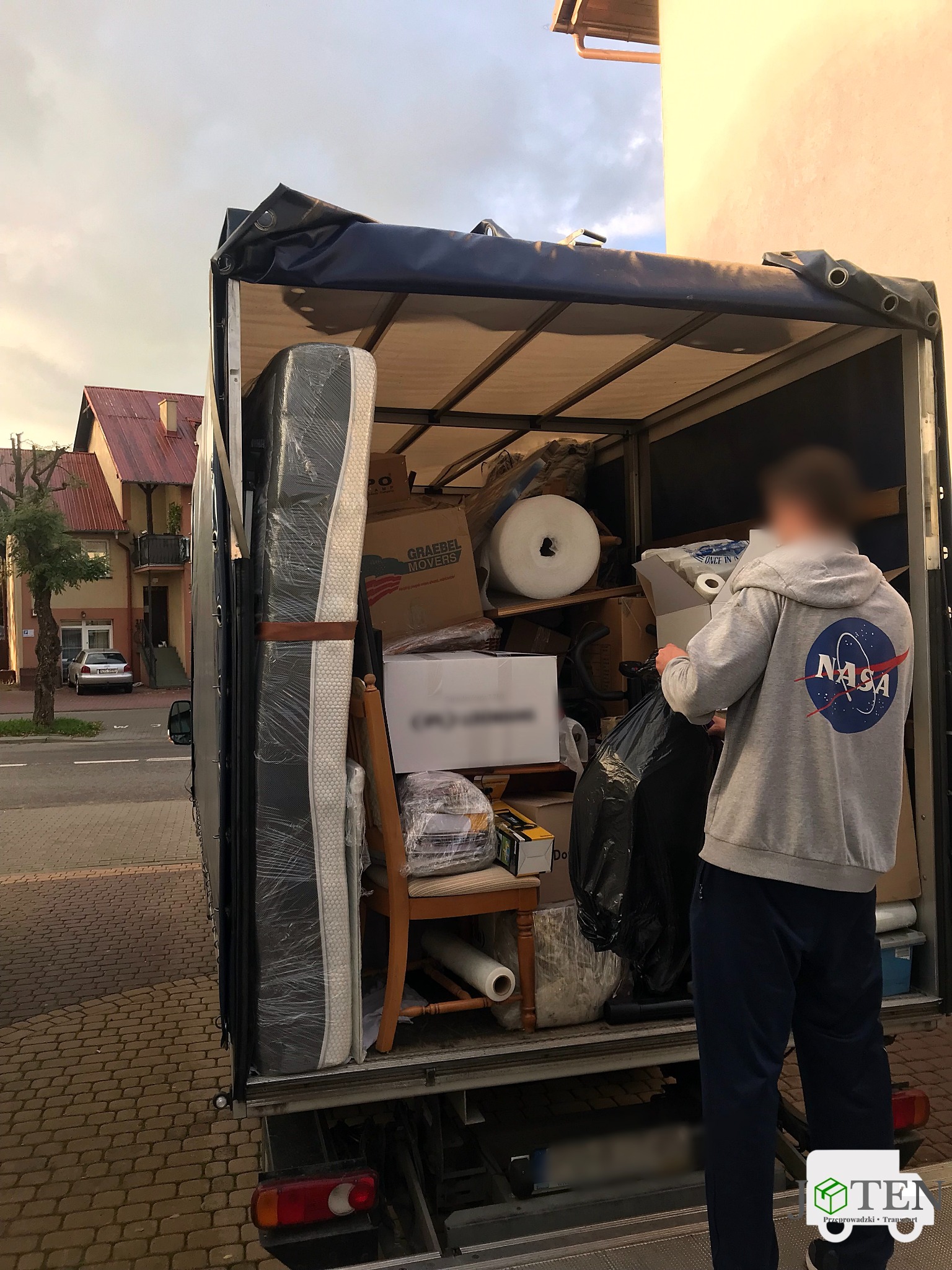 Wnętrze przestrzeni ładunkowej wypełnione kartonami, meblami i materacem, z widoczną osobą w bluzie NASA wkładającą czarny worek do środka, sugerujące załadunek podczas relokacji.