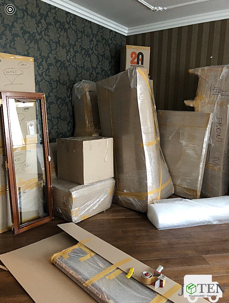 Spakowane meble i przedmioty w folii i kartonach podczas przygotowań do transportu; widoczne oznaczenia 'GLASS' na kartonach, brązowa podłoga i fragment tapety w tle.