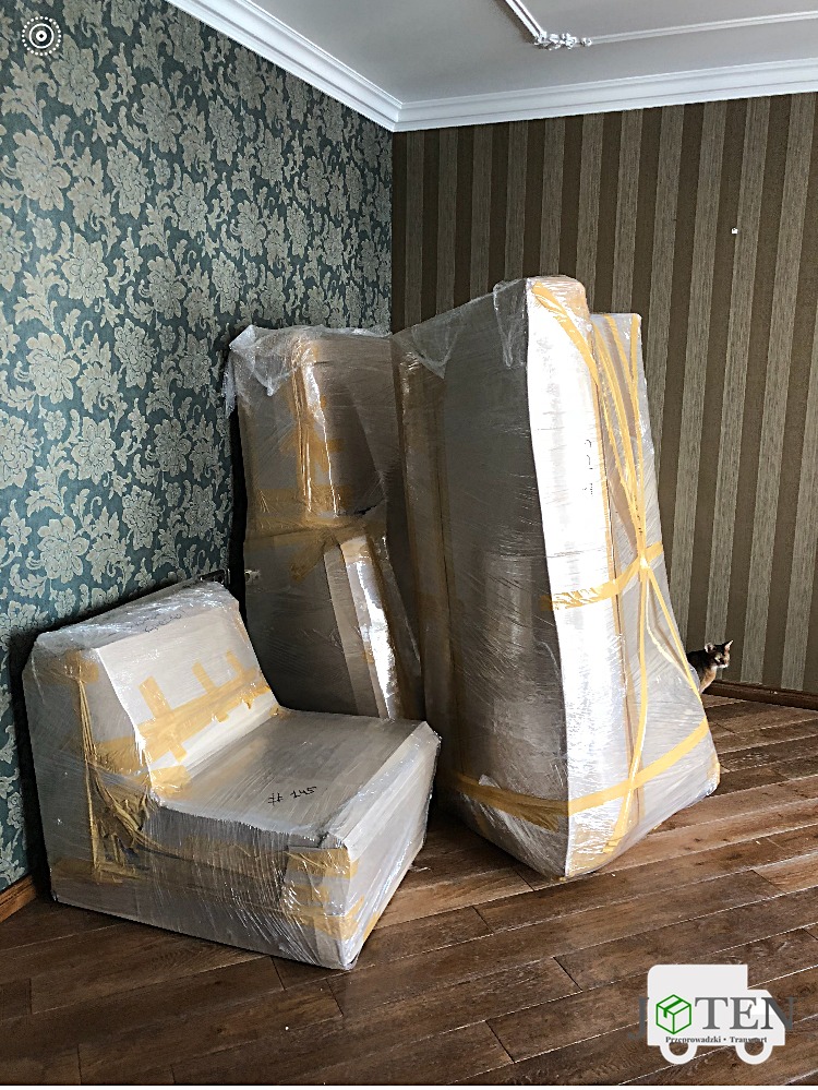 Meble owinięte folią i zabezpieczone taśmą, przygotowane do transportu w pokoju z tapetami w kwiaty i brązowe pasy, z ukrytym kotem w tle.