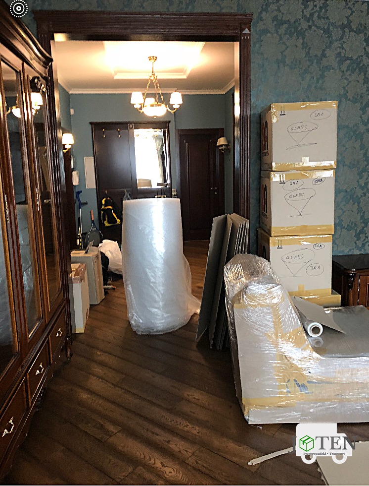 Wnętrze mieszkania z przygotowanymi do transportu przedmiotami: kartony z napisem 'GLASS', meble owinięte folią bąbelkową i stretch, panele zabezpieczone kartonem, widoczny parkiet i drzwi w tle.