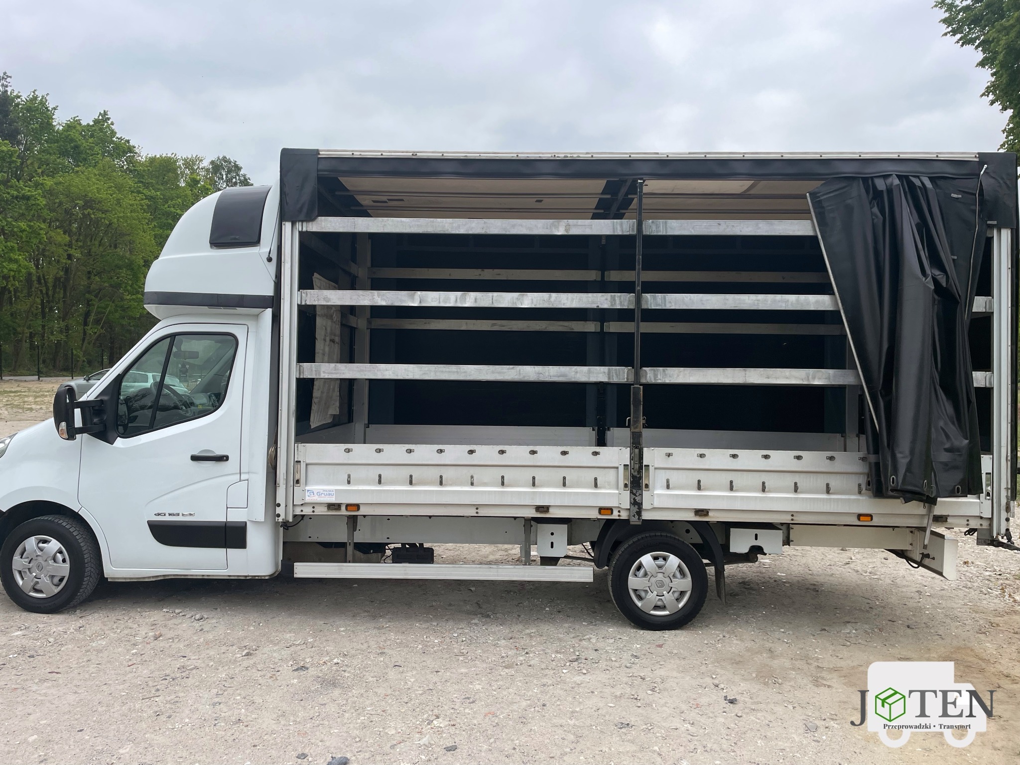Biały samochód dostawczy z otwartą przestrzenią ładunkową i częściowo odsłoniętą plandeką, widok z boku na szutrowym podłożu.