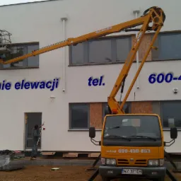 Ekipa maluje elewację budynku biurowego z użyciem podnośnika koszowego i wałka na długim kiju, widoczny samochód z numerem telefonu firmy.