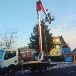 Podnośnik koszowy na samochodzie ciężarowym, wysięgnik uniesiony do góry, obok słupa energetycznego, w tle budynki i drzewa w zimowej scenerii.