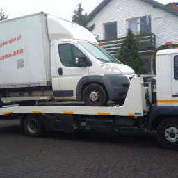 Laweta z białym busem na platformie, zaparkowana na brukowanej drodze przed domem z białymi ścianami i brązowym dachem. Na bucie widoczne logo i numer telefonu firmy.