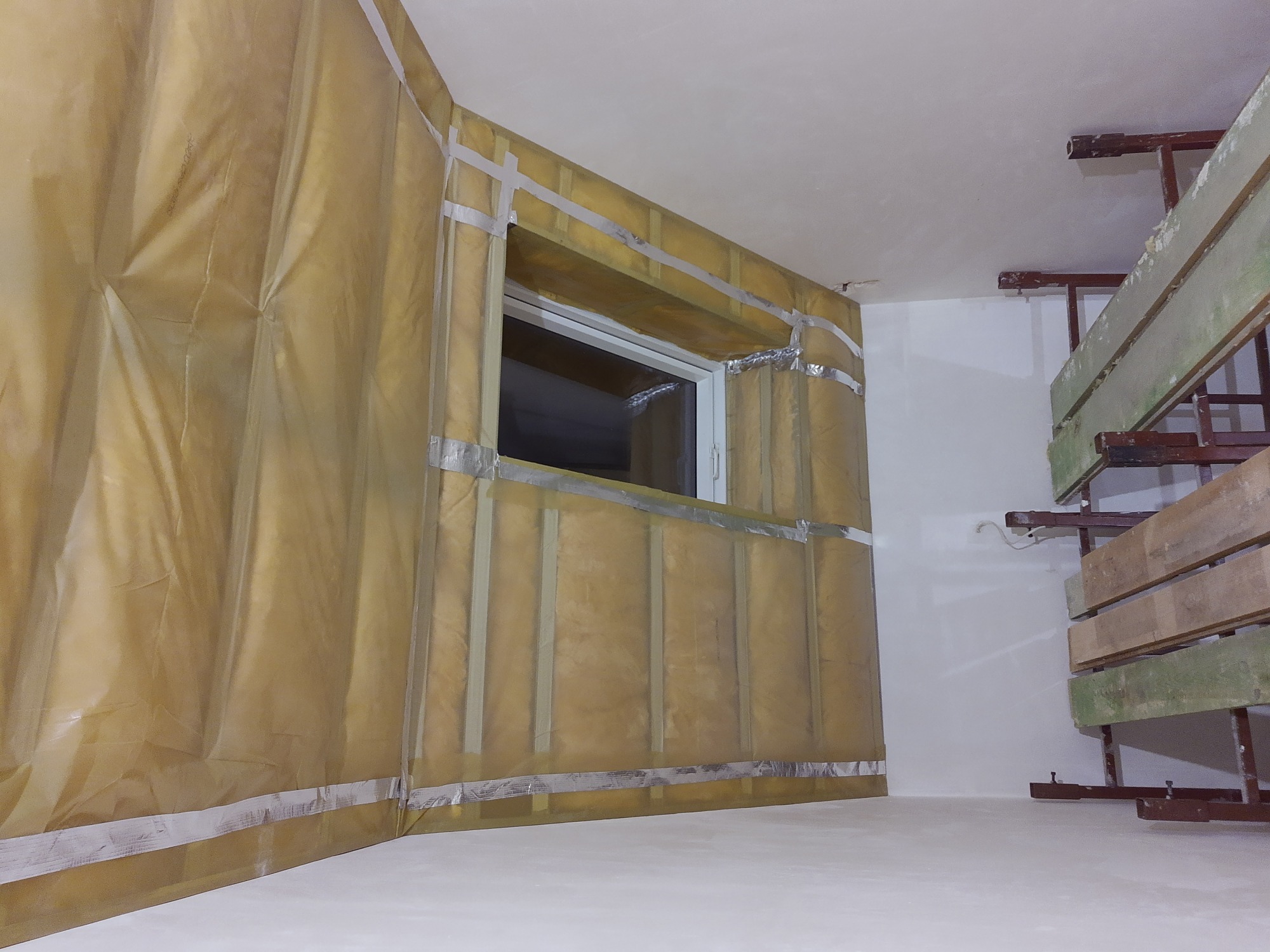 Izolacja termiczna ściany z oknem, zabezpieczona folią i taśmą aluminiową, widoczny fragment rusztowania w rogu pomieszczenia.