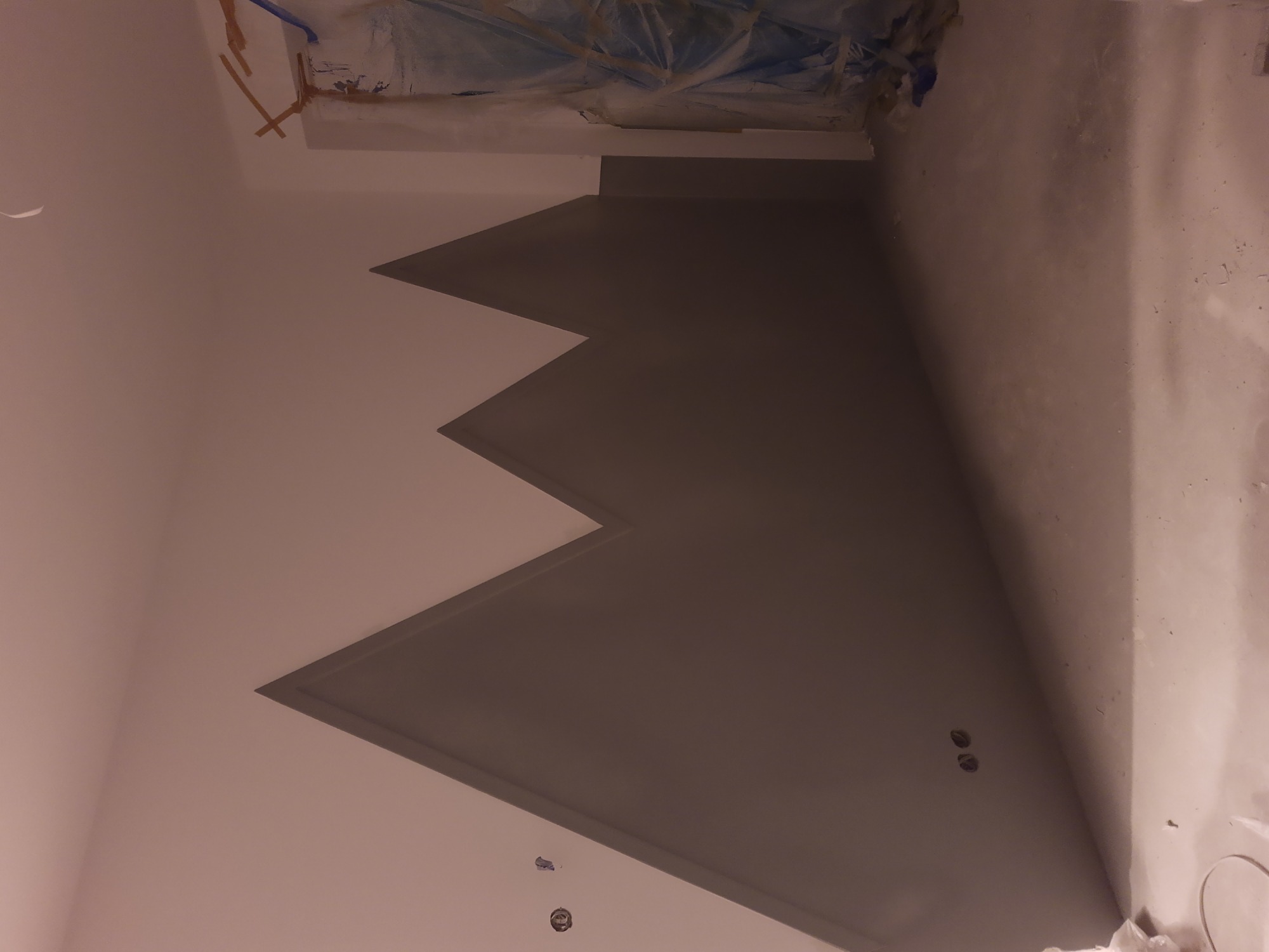 Ściana z nowo pomalowaną, szarą, trójkątną dekoracją. Widoczne niedoskonałości w wykończeniu, zakryte folią elementy oraz otwory na gniazdka elektryczne.