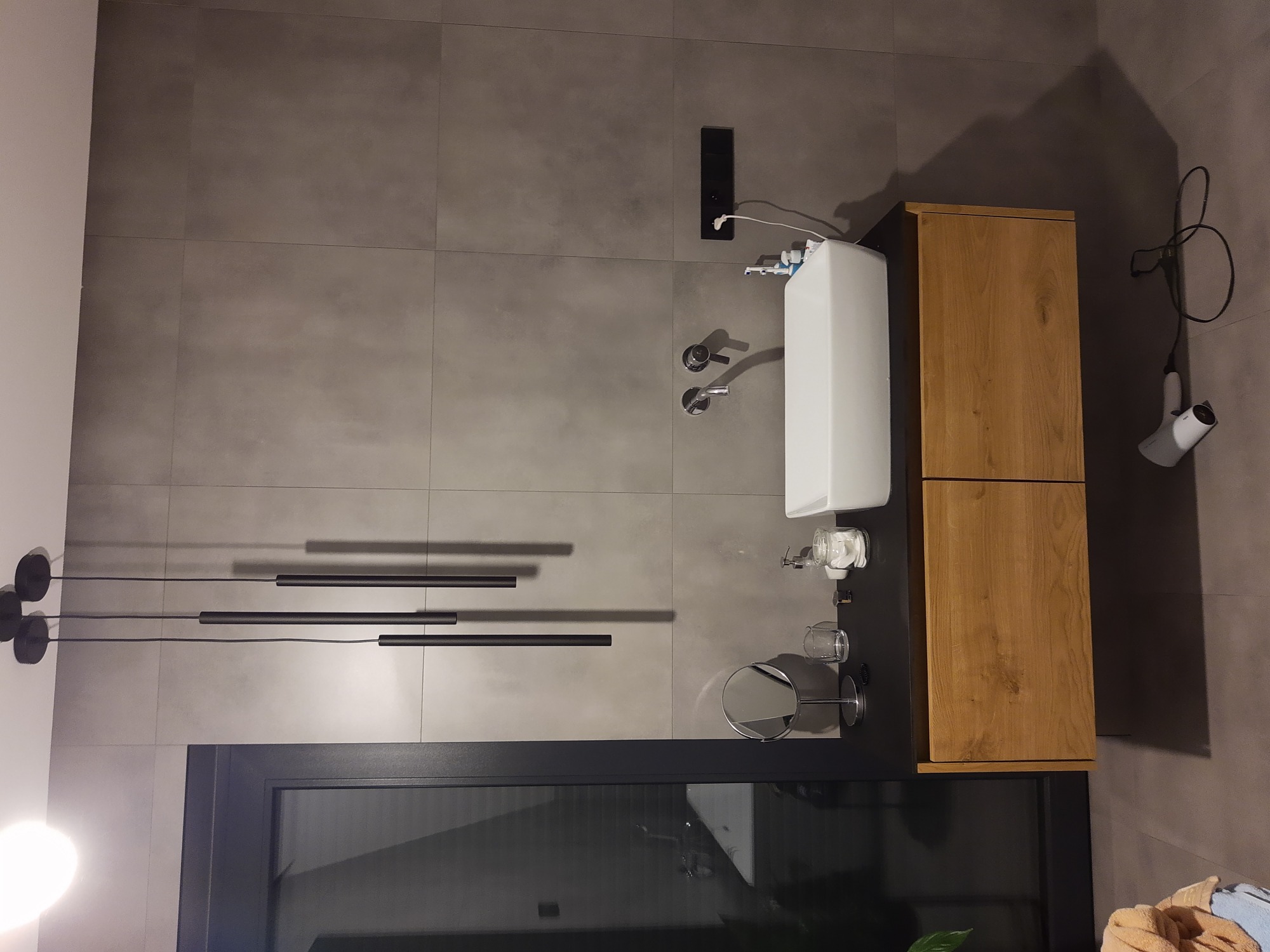 Nowoczesna łazienka z szarymi płytkami, wiszącą szafką z drewnianym frontem, białą umywalką, czarną armaturą i podłużną lampą sufitową.