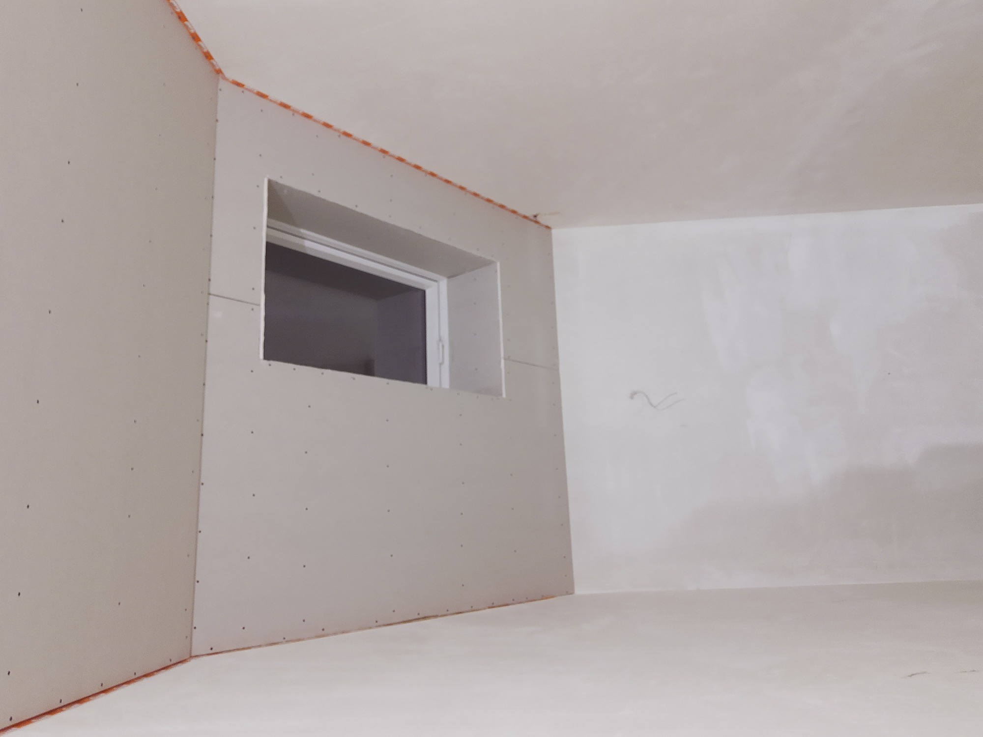 Pokój w trakcie przygotowania do malowania: ściany wyłożone płytami gipsowo-kartonowymi, widoczne łączenia i otwór okienny, narożniki zabezpieczone listwami.