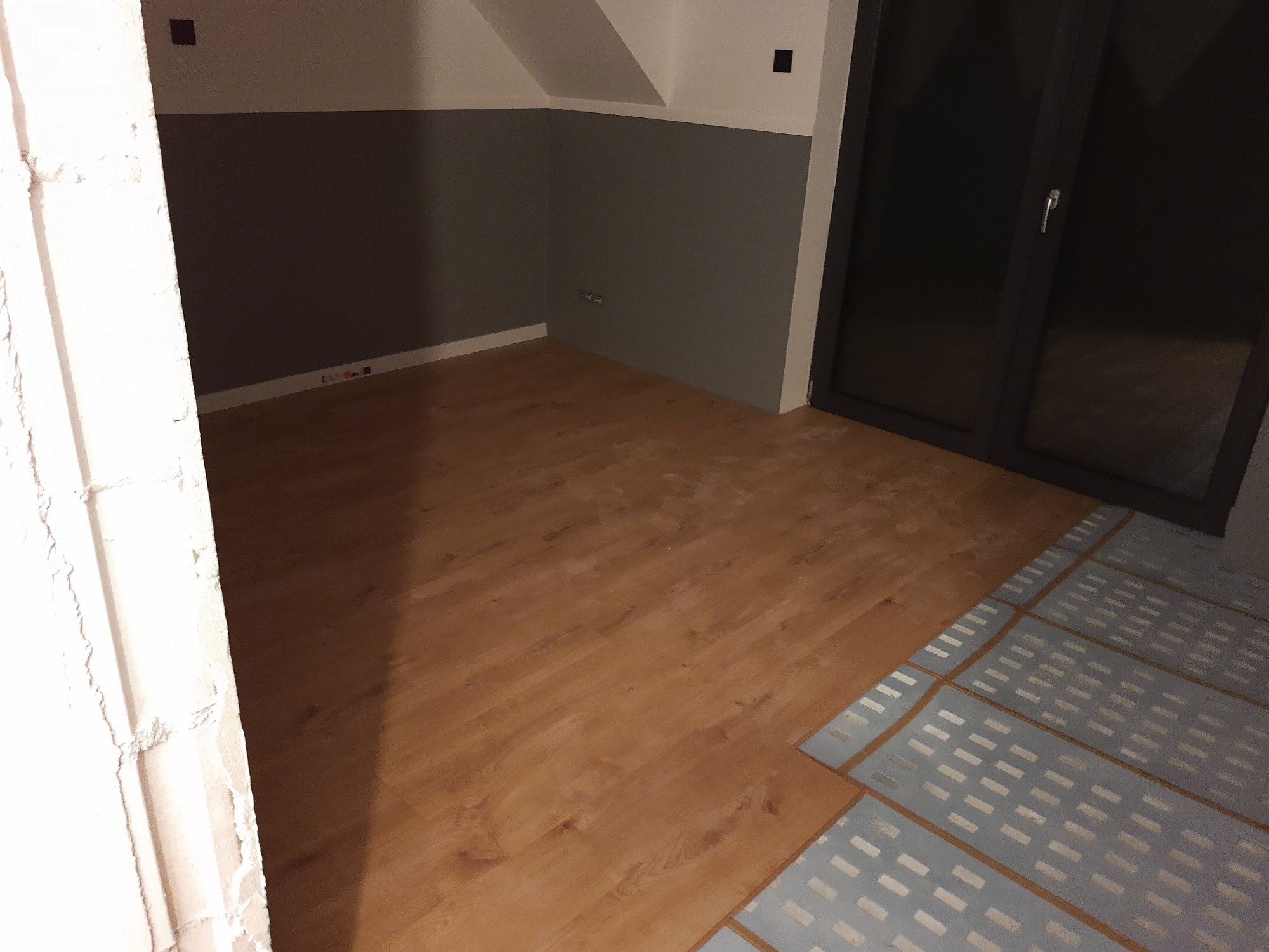 Fragment pokoju w trakcie układania paneli podłogowych; widoczne ułożone panele oraz podkład pod panele, ściany pomalowane dwukolorowo, widoczny również fragment ościeżnicy.