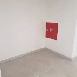 Wnęka w rogu pomieszczenia z szarą podłogą wyłożoną płytkami. Na ścianie zamontowana czerwona szafka hydrantowa z symbolem graficznym. Minimalistyczne wnętrze.