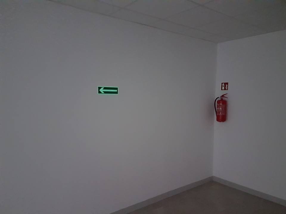 Wewnętrzne ujęcie narożnika pomieszczenia z białymi ścianami, gdzie widoczny jest fluorescencyjny zielony znak kierunkowy oraz czerwona gaśnica z instrukcją obsługi zawieszona na ścianie.