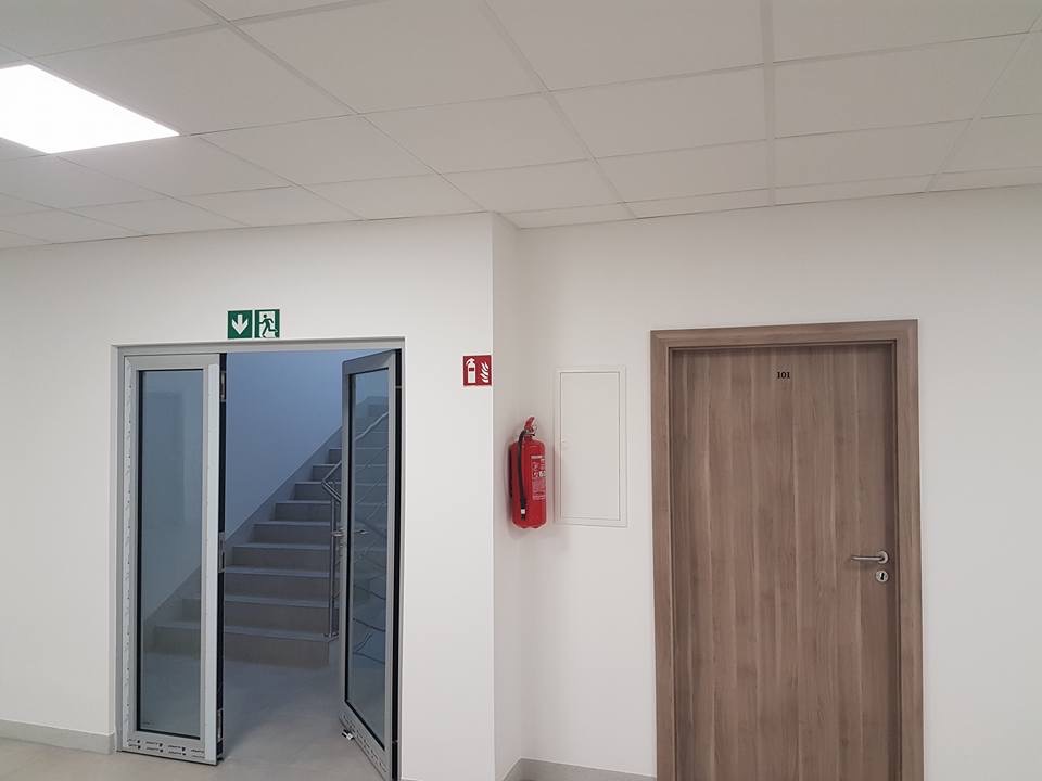 Korytarz biurowy z otwartymi szklanymi drzwiami ewakuacyjnymi prowadzącymi na schody, gaśnicą na ścianie i brązowymi drzwiami z numerem 101.