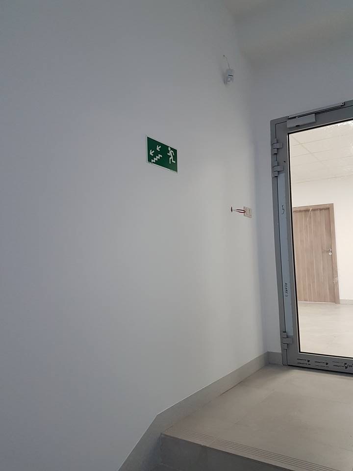 Znak ewakuacyjny z zielonym piktogramem na białej ścianie, widoczny fragment schodów i drzwi z aluminiową ramą.