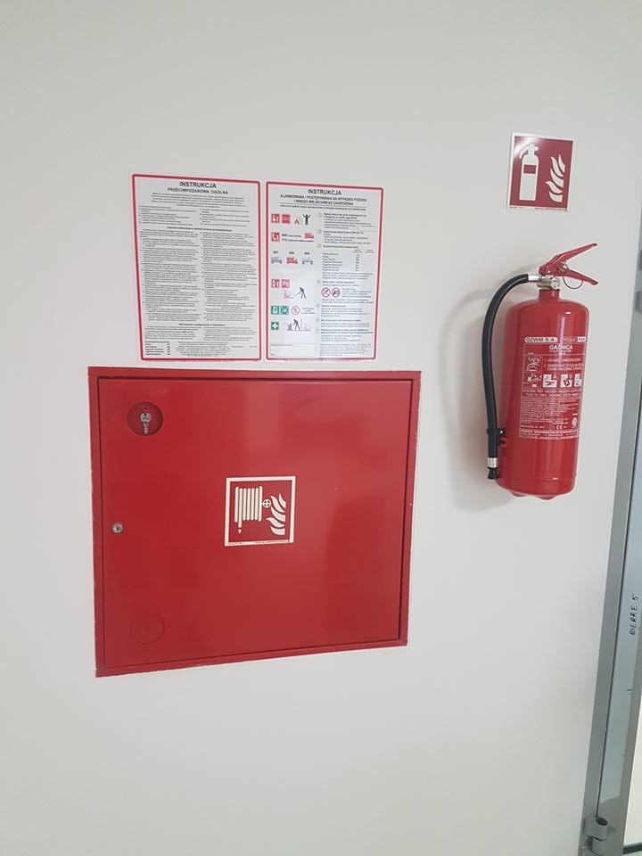 Czerwona szafka hydrantowa z instrukcją, gaśnica i schemat rozmieszczenia sprzętu przeciwpożarowego na białej ścianie.