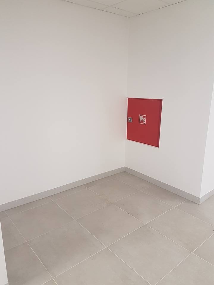 Wnęka w rogu pomieszczenia z szarą podłogą wyłożoną płytkami. Na ścianie zamontowana czerwona szafka hydrantowa z symbolem graficznym. Minimalistyczne wnętrze.