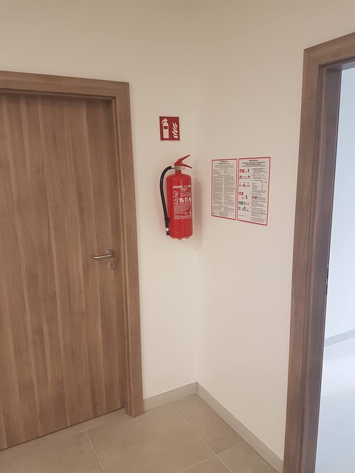 Czerwona gaśnica proszkowa zawieszona na białej ścianie obok piktogramu gaśnicy i instrukcji bezpieczeństwa pożarowego w korytarzu z dwojgiem drewnianych drzwi.