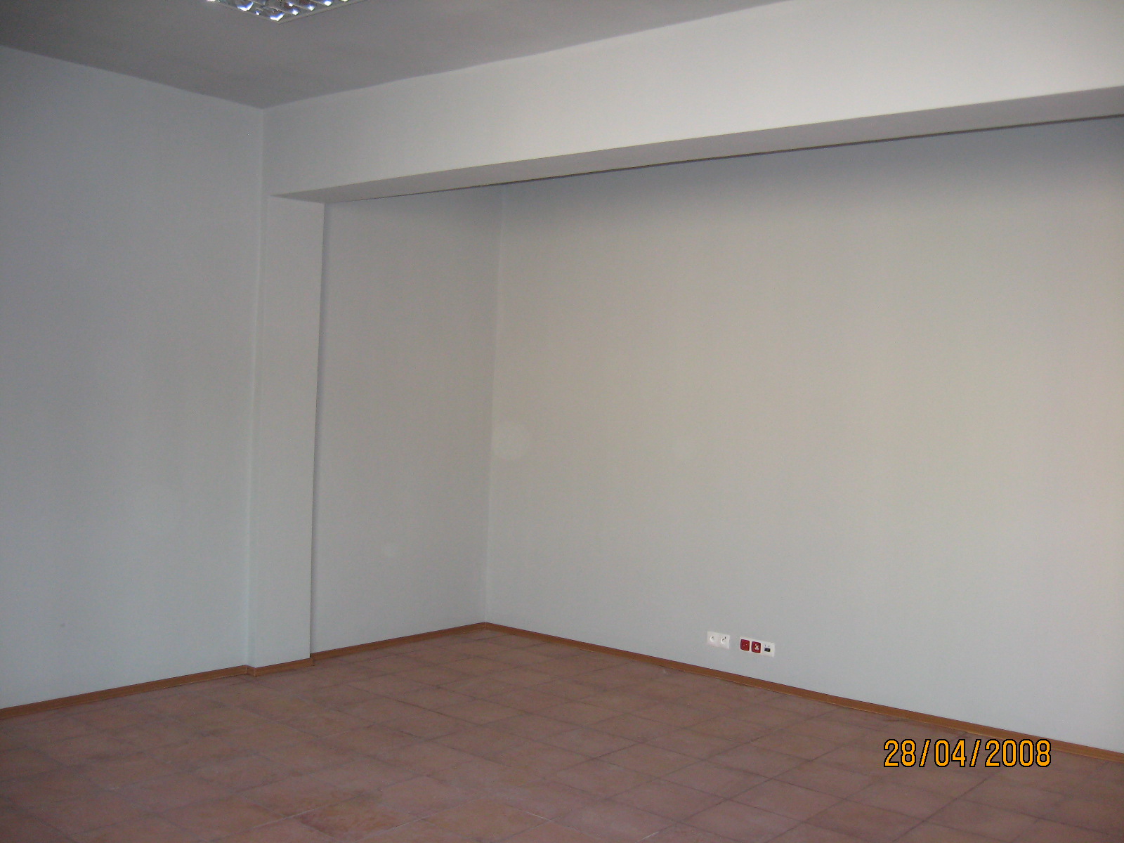 Puste biuro po remoncie z brązową podłogą z płytek, szare ściany i widocznymi gniazdkami elektrycznymi.