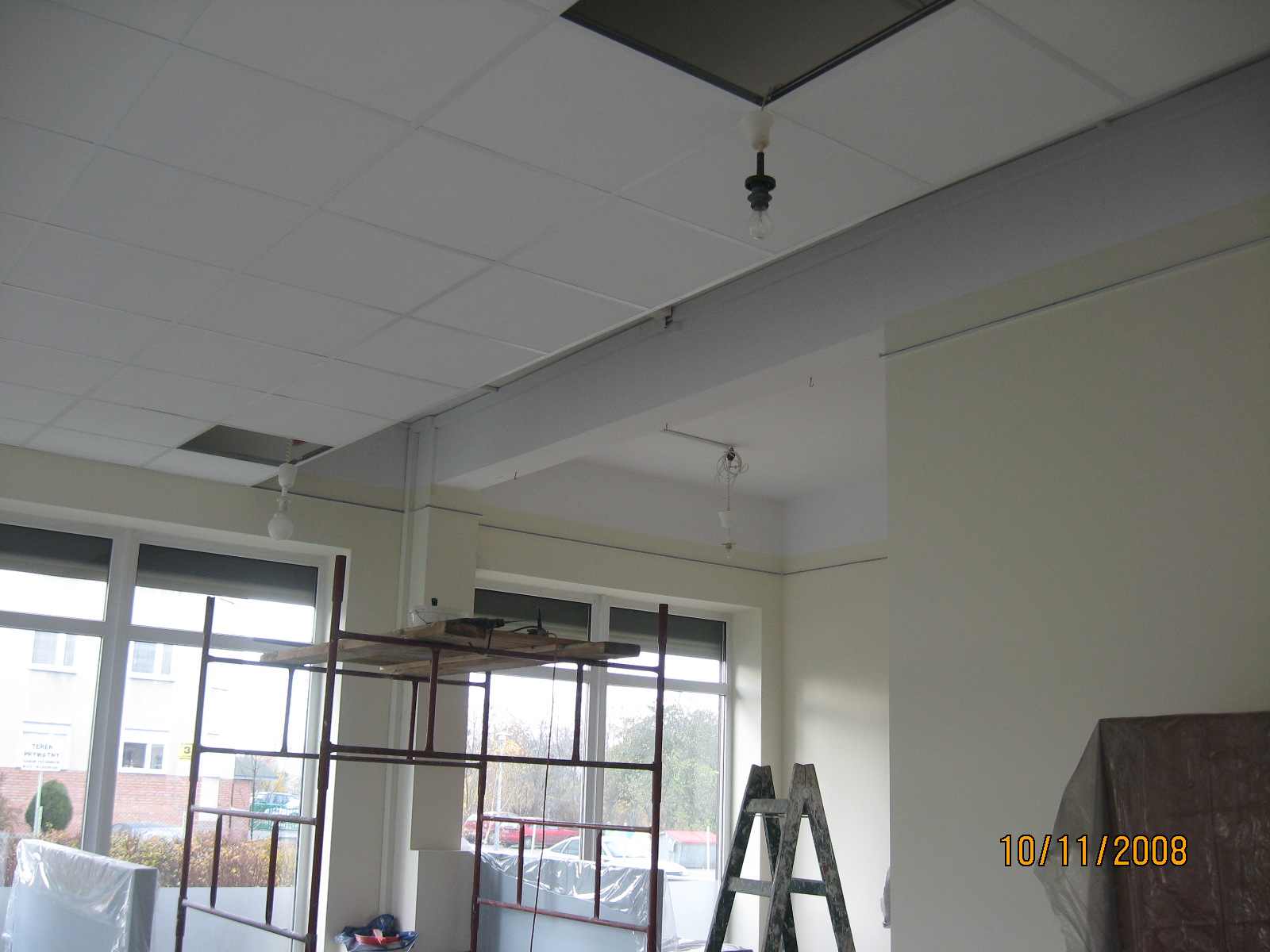 Wnętrze remontowanego biura z widocznym rusztowaniem, drabina oparta o ścianę, podwieszany sufit z otwartymi panelami i wiszącymi żarówkami na kablach.