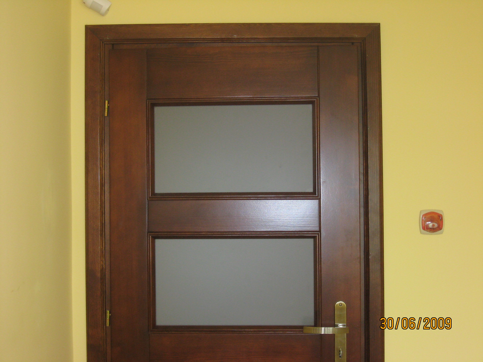 Ciemnobrązowe, przeszklone drzwi wewnętrzne z złotą klamką na żółtej ścianie z datą 30/06/2009 w prawym dolnym rogu.