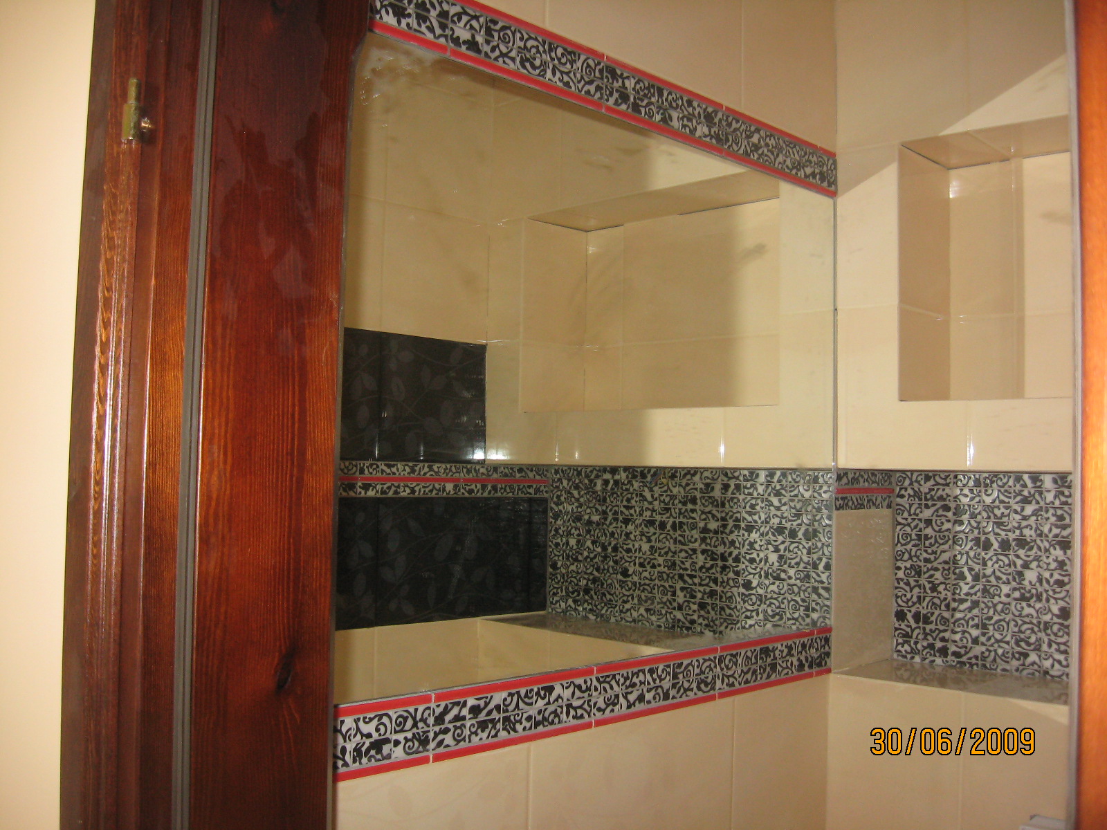 Wykończona łazienka z beżowymi i czarnymi płytkami, dekoracyjnym bordiurem i wnękami, widoczne fragmenty drewnianej ościeżnicy drzwi